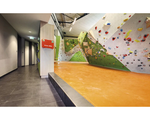 Innenansicht einer Kletterhalle mit Kletterwänden, bunten Klettergriffen und einem Fallschutzboden sowie einem Erste Hilfe Kasten an der Wand.