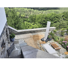 Une terrasse en pierre naturelle avec des meubles de jardin en bois, un parasol et une vue sur la forêt pendant des travaux avec du sable au sol.