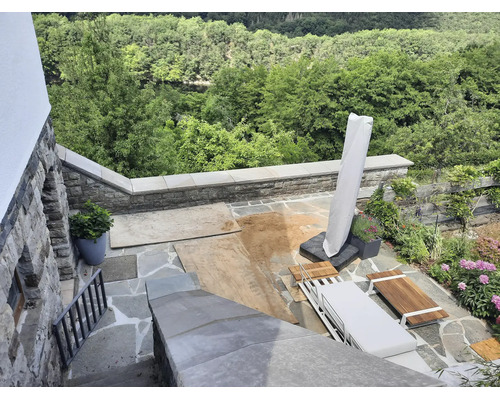 Une terrasse en pierre naturelle avec des meubles de jardin en bois, un parasol et une vue sur la forêt pendant des travaux avec du sable au sol.