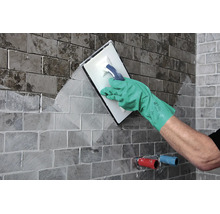 Artisan jointoyant des carreaux sur un mur avec une taloche à joints et des gants de protection