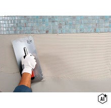 Une main gantée applique de la colle à carrelage sur un mur sous des carreaux de mosaïque à l'aide d'une taloche crantée. (Image AI modified or generated)