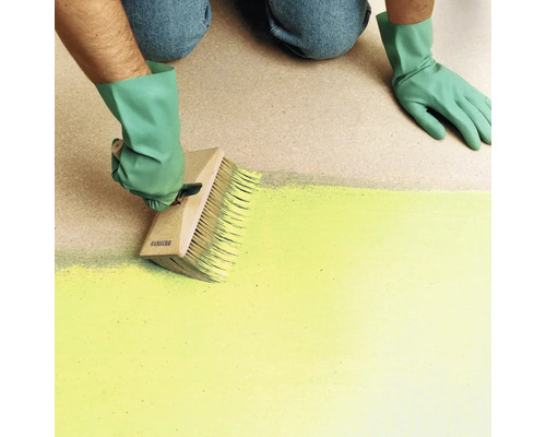 Une personne portant des gants applique de la peinture sur une surface avec une grande brosse à encoller en bois.