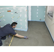 Un artisan lisse une chape fraîchement posée avec une taloche dans une pièce en rénovation avec des cloisons en plaques de plâtre.