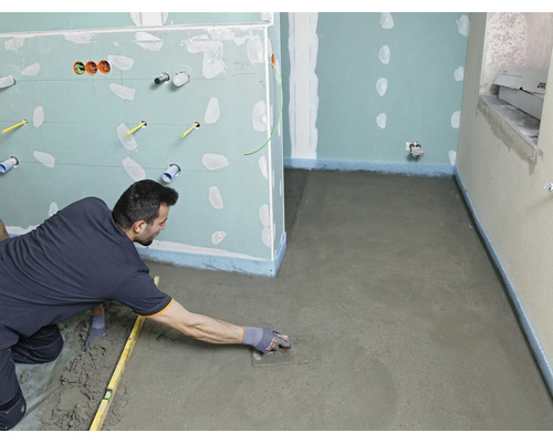 Un artisan lisse une chape fraîchement posée avec une taloche dans une pièce en rénovation avec des cloisons en plaques de plâtre.