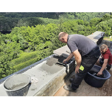 Zwei Handwerker mauern eine Steinmauer im Außenbereich und verstreichen Mörtel mit einer Maurerkelle vor einer grünen Waldkulisse.