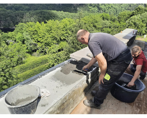 Zwei Handwerker mauern eine Steinmauer im Außenbereich und verstreichen Mörtel mit einer Maurerkelle vor einer grünen Waldkulisse.