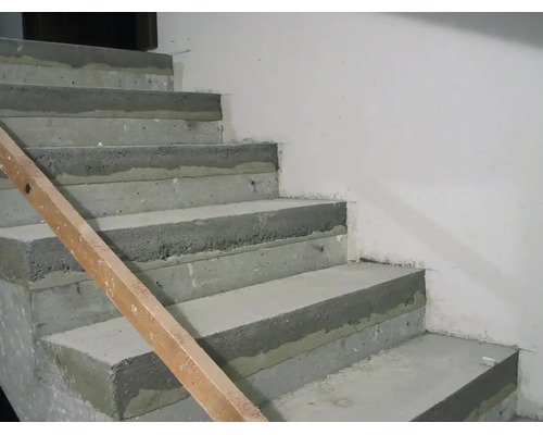 Eine unfertige Betontreppe im Rohbau mit einer hölzernen Führungsschiene an der Seite vor einer glatten Wand.