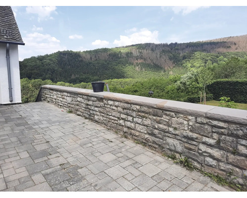 Natursteinmauer aus Bruchsteinen als Terrassenbegrenzung mit Abdeckplatten vor einer bewaldeten Hügellandschaft