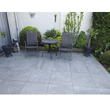 Une terrasse avec des dalles de pierre grand format, des chaises de jardin et une table ronde devant un mur de briques clair avec des plantes.