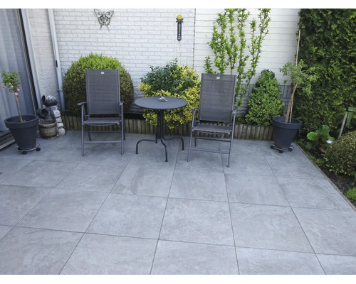 Une terrasse avec des dalles de pierre grand format, des chaises de jardin et une table ronde devant un mur de briques clair avec des plantes.