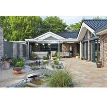Terrasse bien entretenue avec sol en pierre naturelle, petit bassin de jardin avec pont, jacuzzi et coin salon couvert devant une maison moderne.