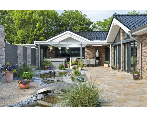 Terrasse bien entretenue avec sol en pierre naturelle, petit bassin de jardin avec pont, jacuzzi et coin salon couvert devant une maison moderne.