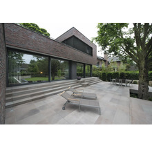 Terrasse moderne avec grandes dalles, chaises longues et coin repas devant une maison en brique avec baies vitrées et jardin.