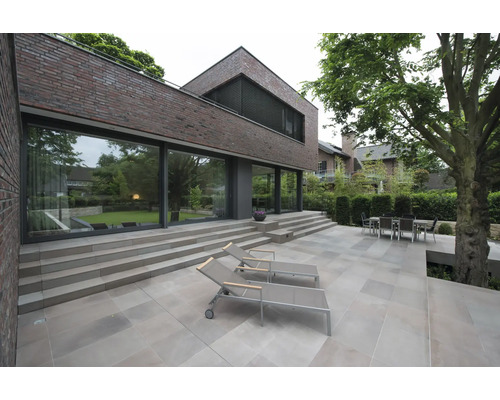 Terrasse moderne avec grandes dalles, chaises longues et coin repas devant une maison en brique avec baies vitrées et jardin.