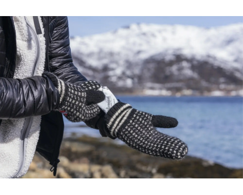 Une personne portant des moufles en tricot insère un chauffe-mains Thermopad dans son gant devant un paysage hivernal avec montagne et lac.