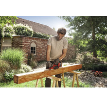 Un homme utilise un rabot sans fil Einhell pour travailler une poutre en bois sur un tréteau dans un jardin ensoleillé.