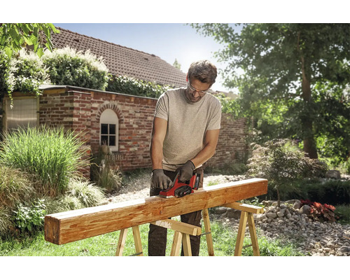 Un homme utilise un rabot sans fil Einhell pour travailler une poutre en bois sur un tréteau dans un jardin ensoleillé.