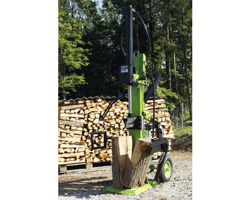 Une fendueuse de bûches verticale se trouve devant un tas de bois sur un chemin forestier.