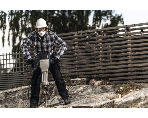 Un homme portant un casque de protection, des lunettes de sécurité et une protection auditive utilise un marteau-piqueur.