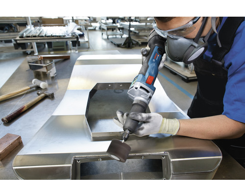 Ein Mann bearbeitet ein Metallstück mit einem Elektrowerkzeug in einer Werkstatt. Persönliche Schutzausrüstung wird getragen.