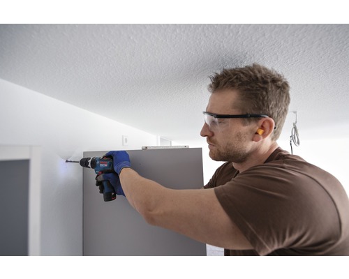 Un homme perce un trou dans un mur avec une perceuse, portant des lunettes de protection et des gants.