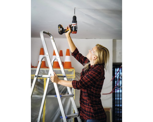 Une femme perce un trou dans le plafond avec une perceuse en se tenant sur une échelle