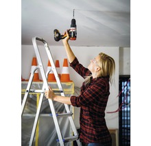 Une femme perce un trou dans le plafond avec une perceuse tout en se tenant sur une échelle.