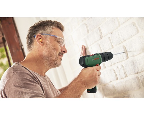 Un homme perce un mur de briques avec une perceuse Bosch, portant des lunettes de protection et des bouchons d''oreille.