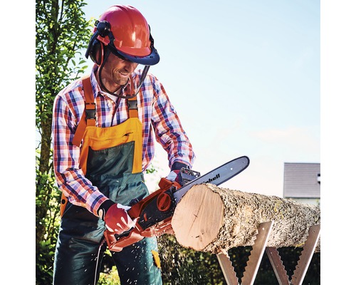 Mann sägt Holzscheit mit einer Elektro Kettensäge auf einem Sägebock, während er einen Helm mit Visier und Gehörschutz trägt