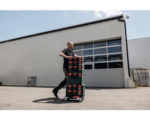 Artisan transportant des boîtes à outils sur une planche à roulettes devant un bâtiment.