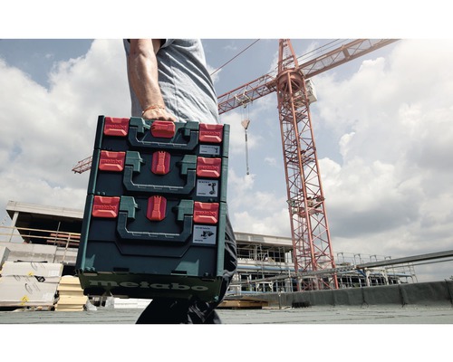 Une personne porte trois coffrets à outils Metabo devant un chantier de construction avec une grue.