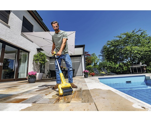 Un homme nettoie une terrasse en pierre avec une brosse de nettoyage de terrasse près d''une piscine.