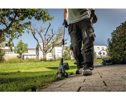 Une personne utilise un coupe-bordure dans le jardin, avec une tondeuse à gazon en arrière-plan.
