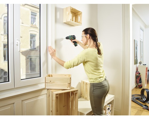 Une femme perce une caisse en bois avec une perceuse sur le mur pour construire une étagère