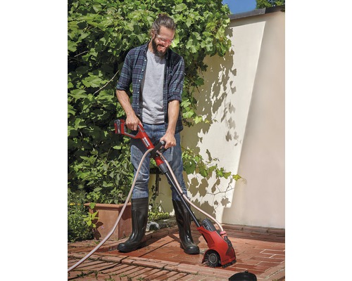 Mann benutzt eine Akku-Fugenbürste im Garten, um Unkraut zwischen Pflastersteinen zu entfernen.