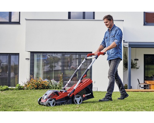 Ein Mann mäht mit einem roten Einhell Akku-Rasenmäher den Rasen vor einem modernen Haus.