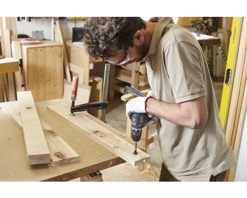 Un artisan perce une planche de bois avec une perceuse sans fil, fixée par un serre-joint.