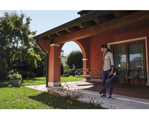 Frau benutzt Laubbläser auf Terrasse, Rasenmäher im Hintergrund