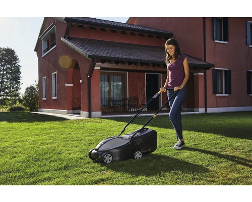 Frau benutzt Rasenmäher im Garten vor Wohnhaus.