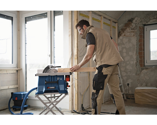 Un homme utilise une scie circulaire de table pour couper une planche de bois. Un aspirateur se trouve à côté.