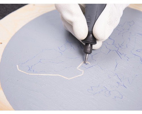 Eine Hand mit Handschuh benutzt ein Multifunktionswerkzeug auf einem bemalten Holzkreis.