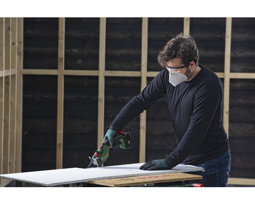 Une personne coupe une plaque de plâtre avec une scie sabre sans fil. Un équipement de sécurité est porté.