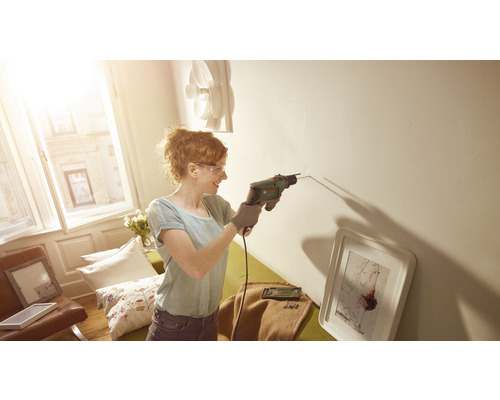 Frau bohrt mit einer Schlagbohrmaschine ein Loch in die Wand in einem hellen Zimmer.