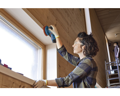 Une femme ponce du bois avec une ponceuse dans une pièce lumineuse.