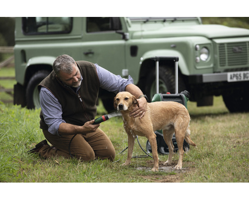 Un homme lave un chien à l'extérieur avec un nettoyeur portable devant un véhicule tout-terrain