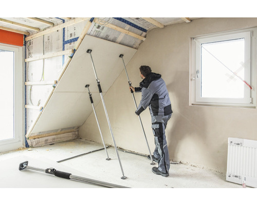 Mann befestigt Gipskartonplatte mit Teleskopstützen an Dachschräge
