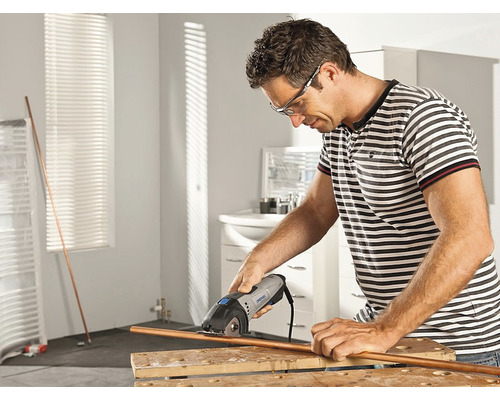 Un homme coupe un tube de cuivre avec un outil Dremel sur un établi dans une salle de bains.