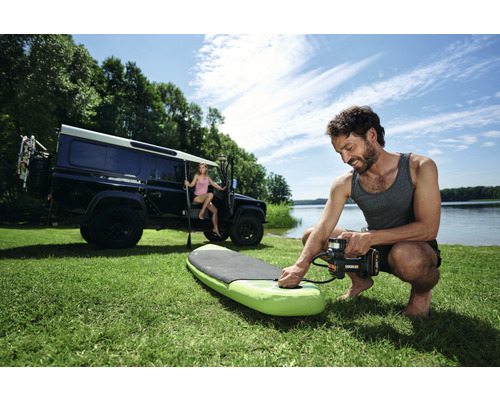 Mann pumpt ein Stand-Up-Paddleboard mit einer elektrischen Pumpe auf dem Rasen auf, im Hintergrund ein Geländewagen und eine Frau