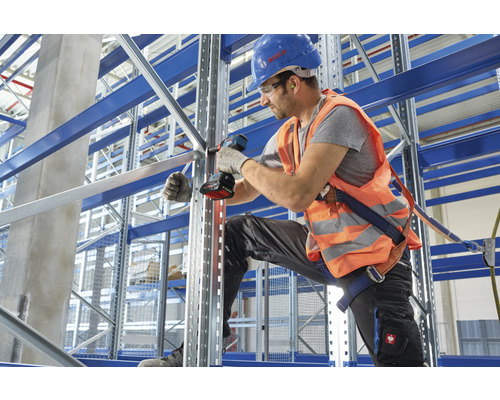 Un homme assemble des rayonnages d'entrepôt avec un tournevis sans fil, un casque de protection et un gilet de sécurité dans un entrepôt