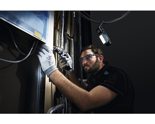 Ein Mann mit Schutzbrille und Handschuhen arbeitet mit einem Akkuschrauber an einem Objekt, beleuchtet von einer Arbeitsleuchte.